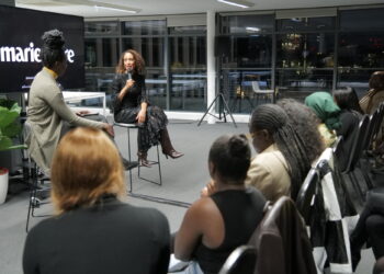 Two women sitting on tall chairs addressing an audience in a room with grey carpet and dark windows
