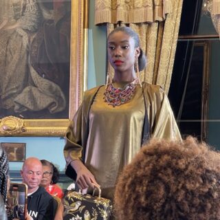 Woman in bronze top and large necklace standing in a period room among spectators