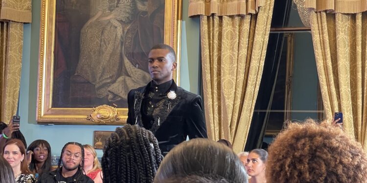 Man in black velevet suit standing in a period room with spectators around