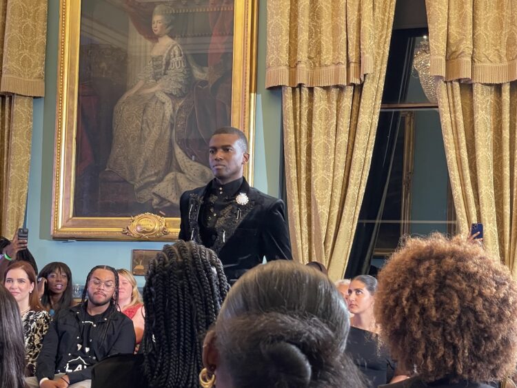 Man in black velevet suit standing in a period room with spectators around